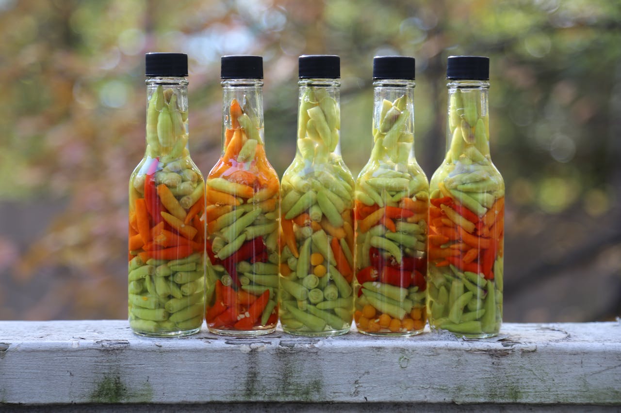 Five bottles filled with vibrant pickled hot peppers displayed outdoors on a sunny day.
