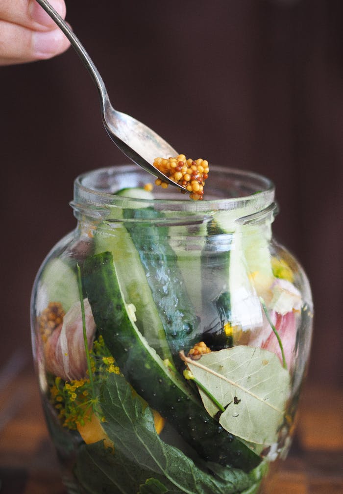 Hand using spoon to add mustard seeds to a jar of fresh pickled vegetables.